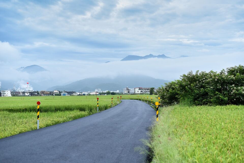 Jalan aspal membelah hamparan hijau dengan barisan gunung di kejauhan.